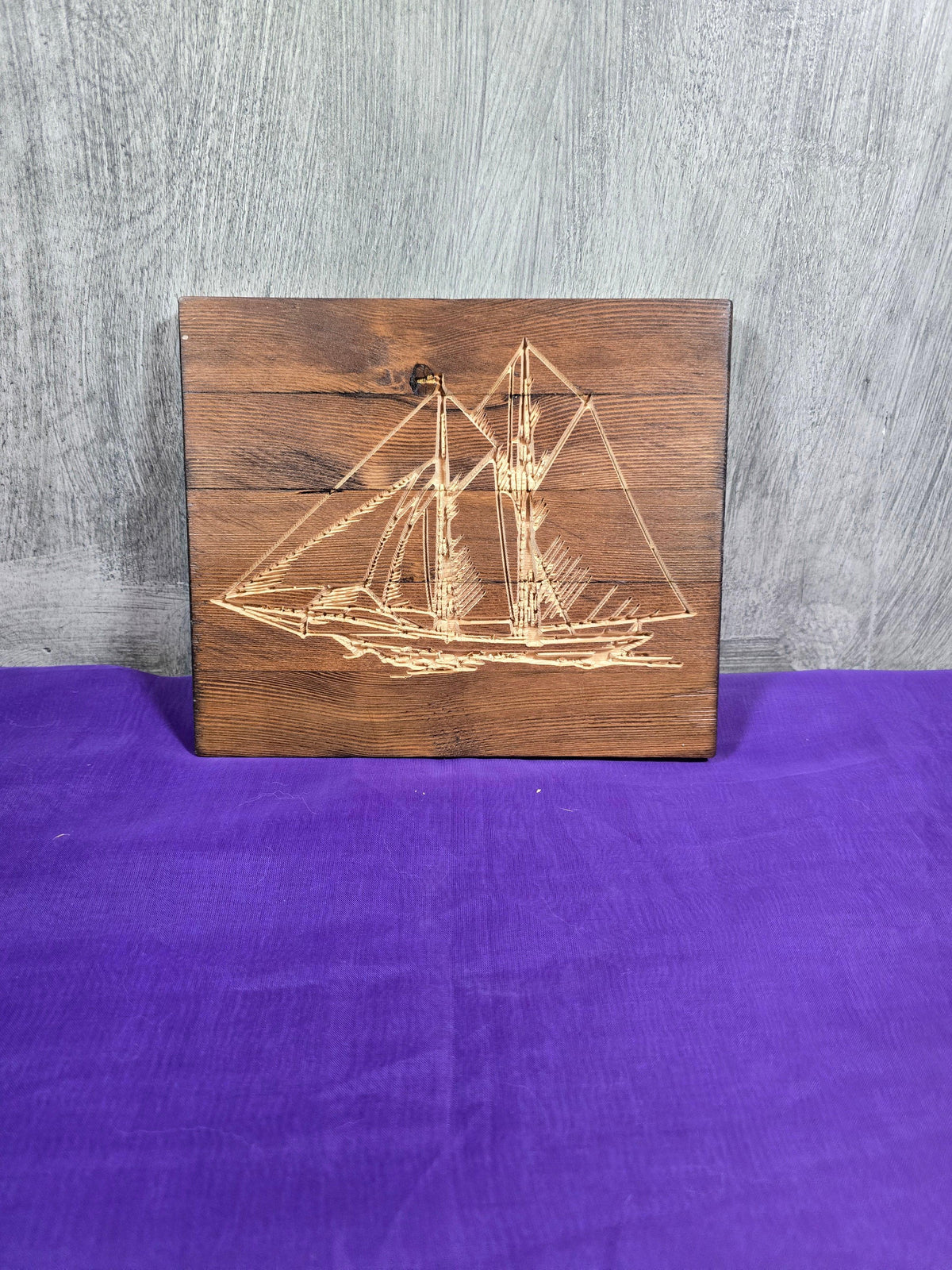 Wooden sailboat engraving on a dark wood plaque, displayed on a purple fabric background.